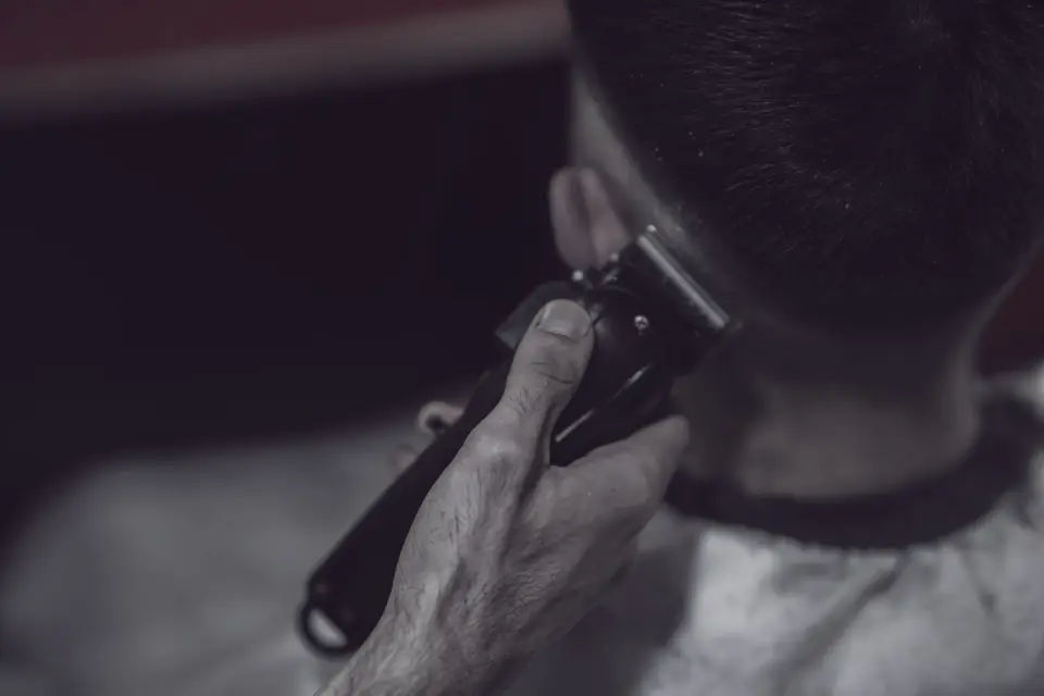 A hand holding a dark clipper to the back of a client’s head, behind the ear