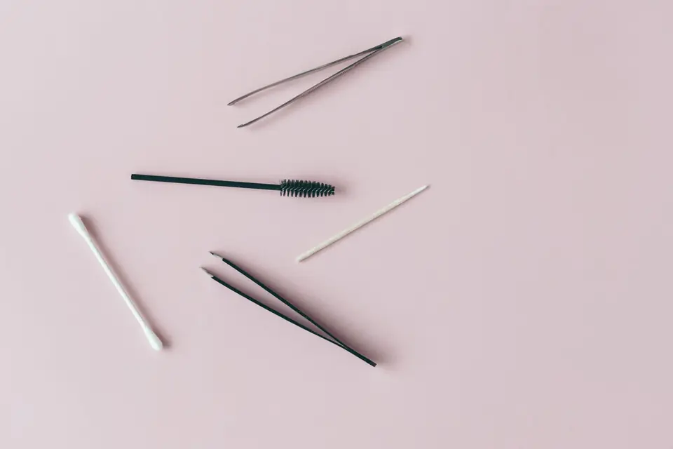 A spoolie, two pairs of tweezers, a cotton bud and a wooden stick on a flat pastel surface
