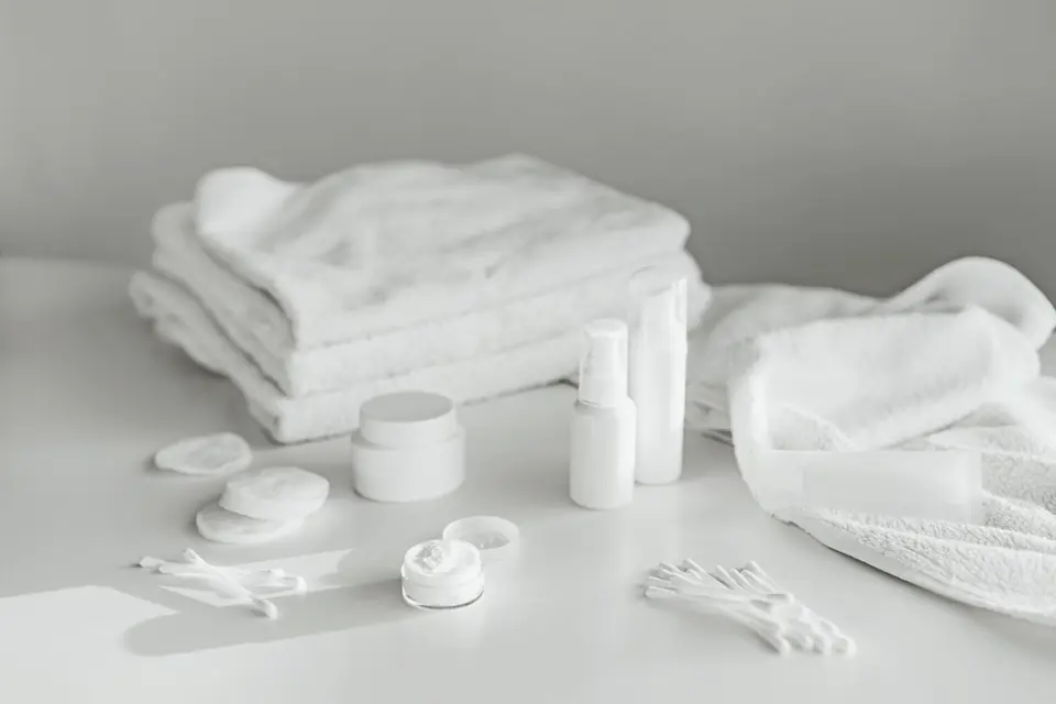 Unlabelled white bottles, a jar, cotton pads and cotton buds beside folded white towels